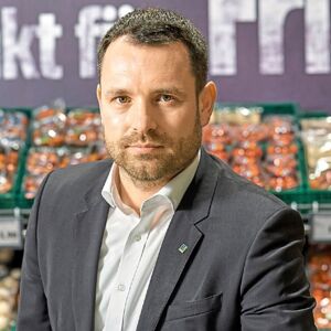 Porträtfoto von Jens Reimer, Geschäftsführer EDEKA Jens, im Hintergrund ein Gemüseregal im EDEKA Markt. Er trägt ein dunkelgraues Sakko und ein weißes Hemd.